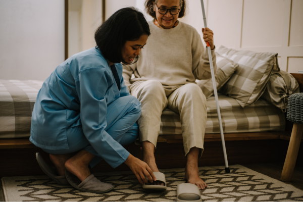 Caregiver helping elderly woman with slippers beside a bed.