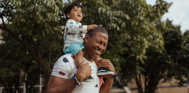 Man carrying a smiling child on his shoulders outdoors, surrounded by trees.